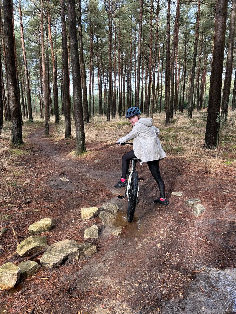 Teenage girl in a helmet pushing a mountain bike along a muddy forest trail.