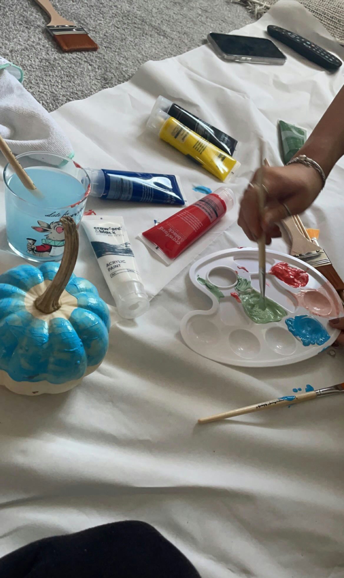 Hand painting a blue pumpkin with a paintbrush, surrounded by paint tubes, a palette and brushes.