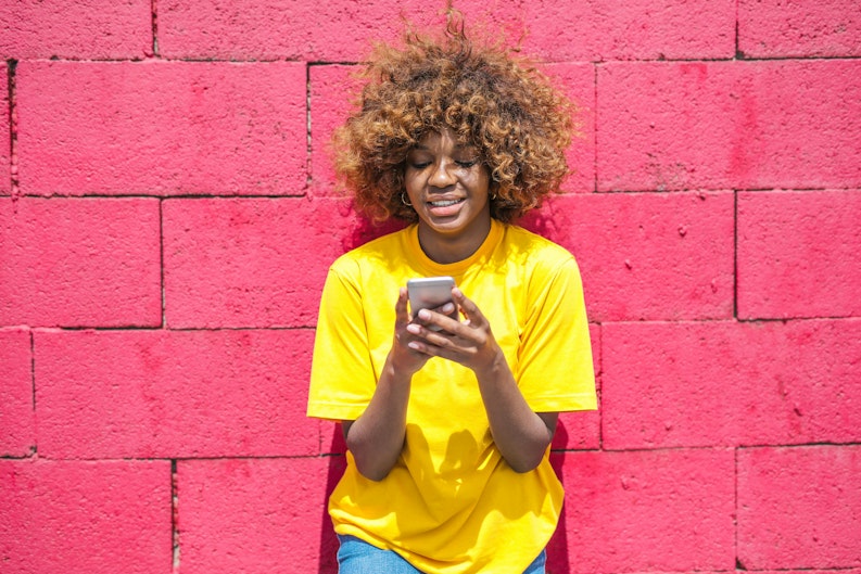 Teenage girl in a yellow T-shirt looking at her phone against a pink brick wall.