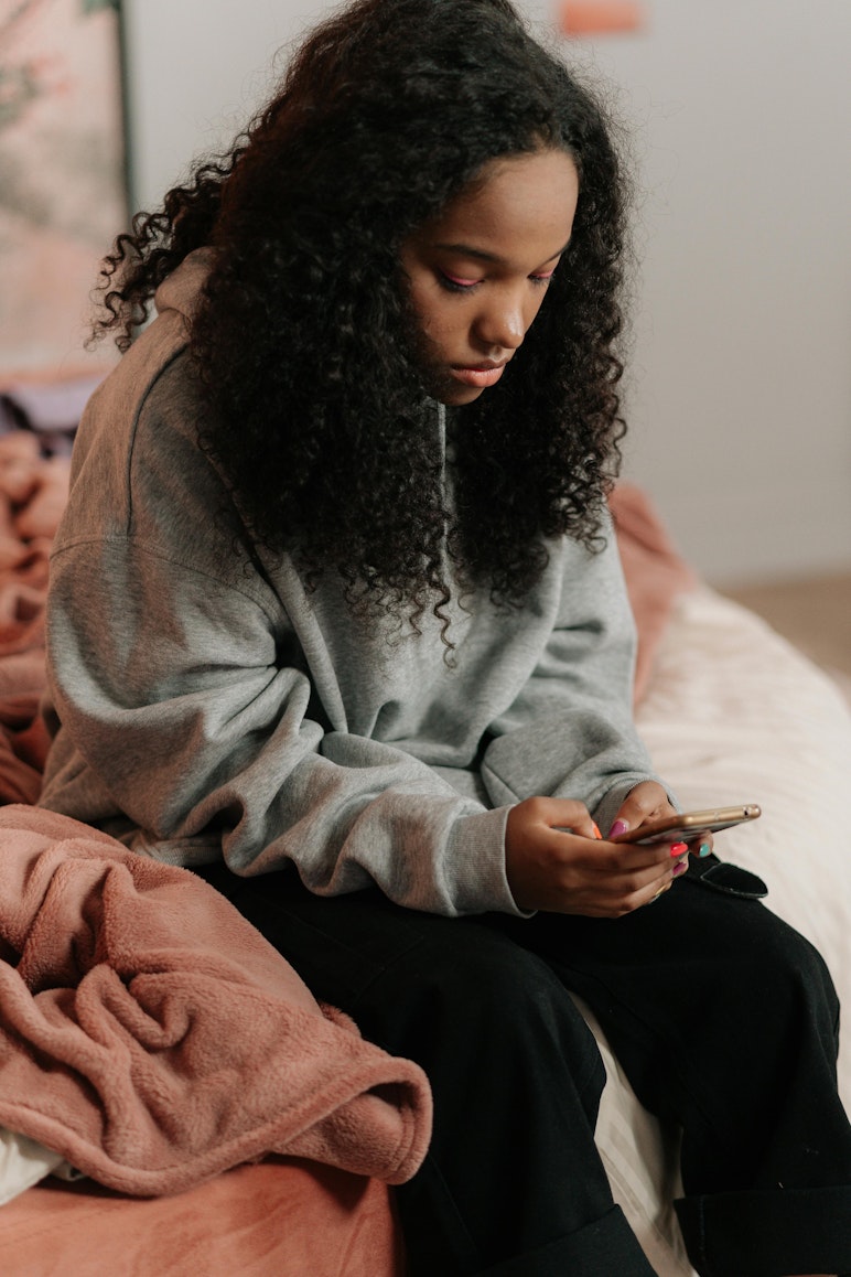 Teenage girl sitting on a bed looking down at her phone.
