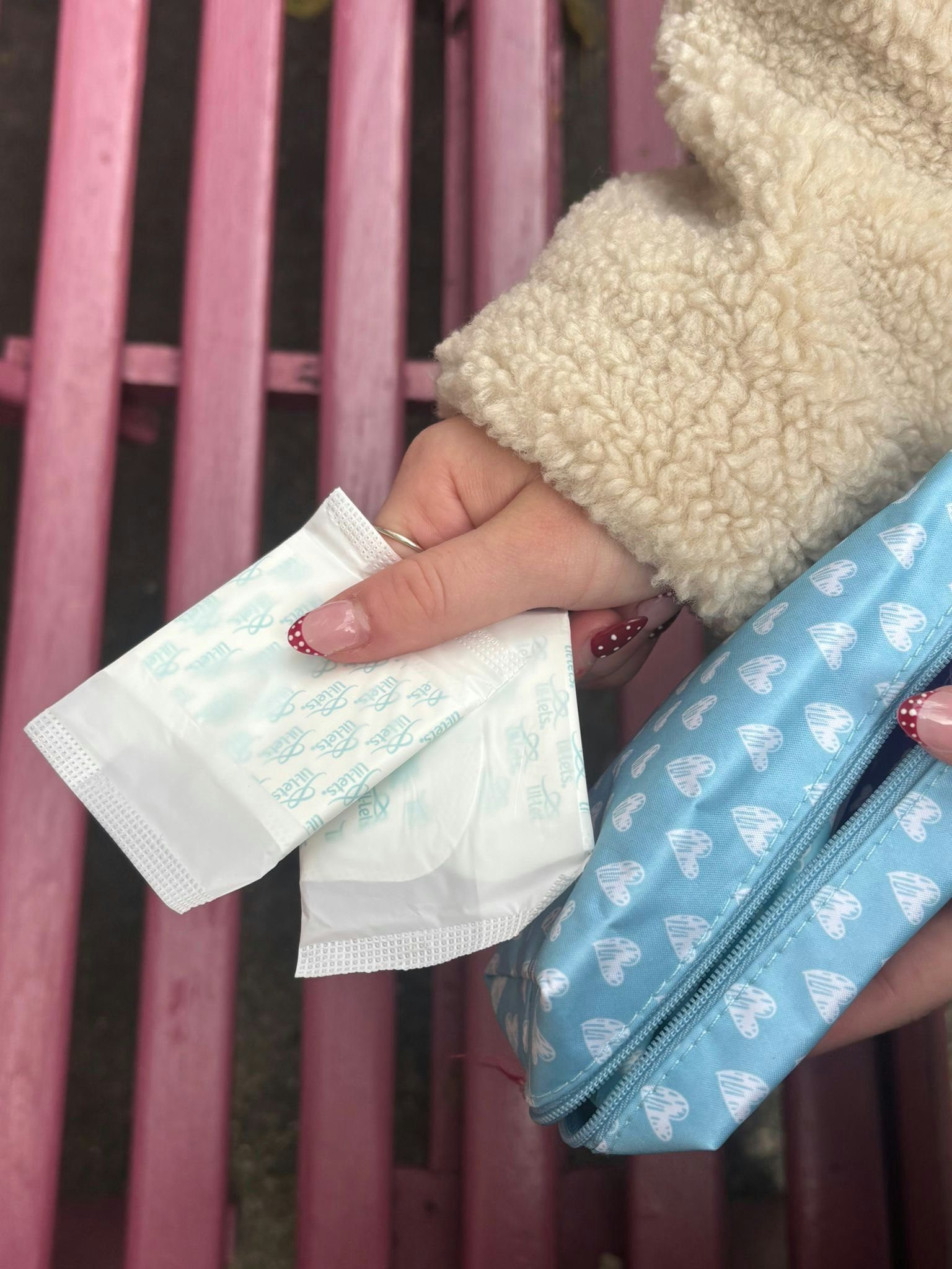 Hand holding two wrapped period pads next to a light blue heart-print pouch on a pink bench.