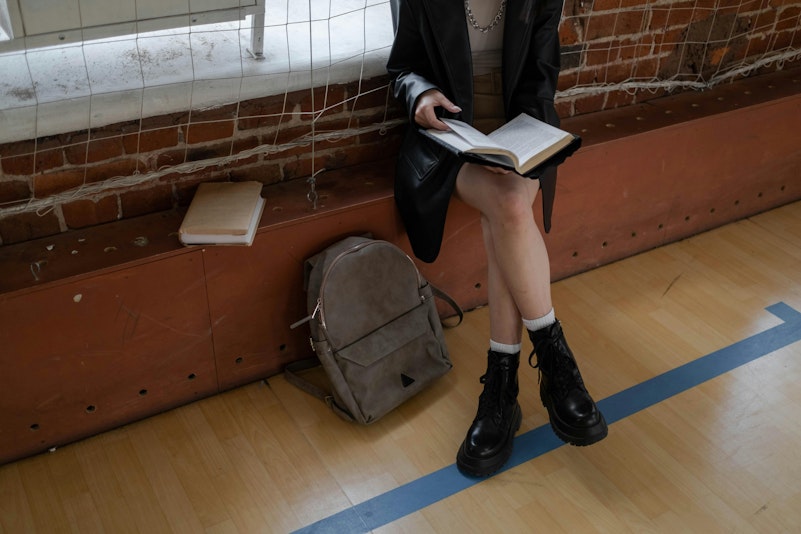 Teenage girl in school uniform sitting on a bench reading a book, with a backpack on the floor beside her.
