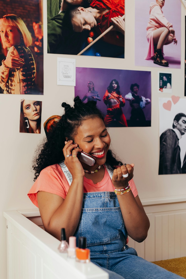 Teenage girl laughing while on a phone call, sitting in front of a wall covered in magazine cutouts and photos.