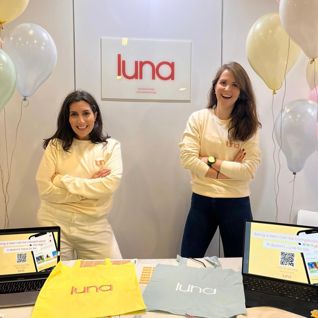 Two women standing together at a luna branded stand with balloons, tote bags and laptops.