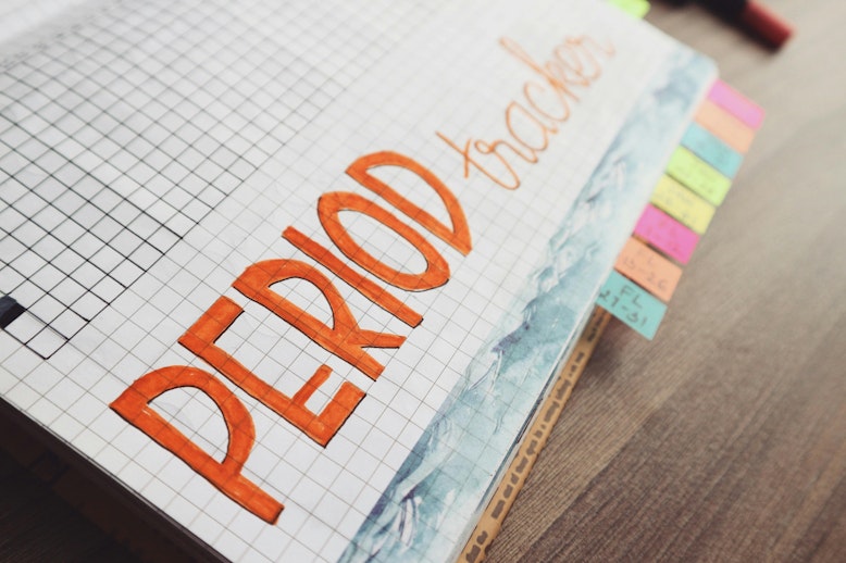 Notebook page with "period tracker" hand-lettered in orange bubble text on grid paper, with colourful sticky tabs on the side.
