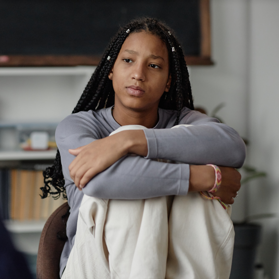 Teenage girl sitting with her arms wrapped around her knees, looking off to the side with a thoughtful expression.