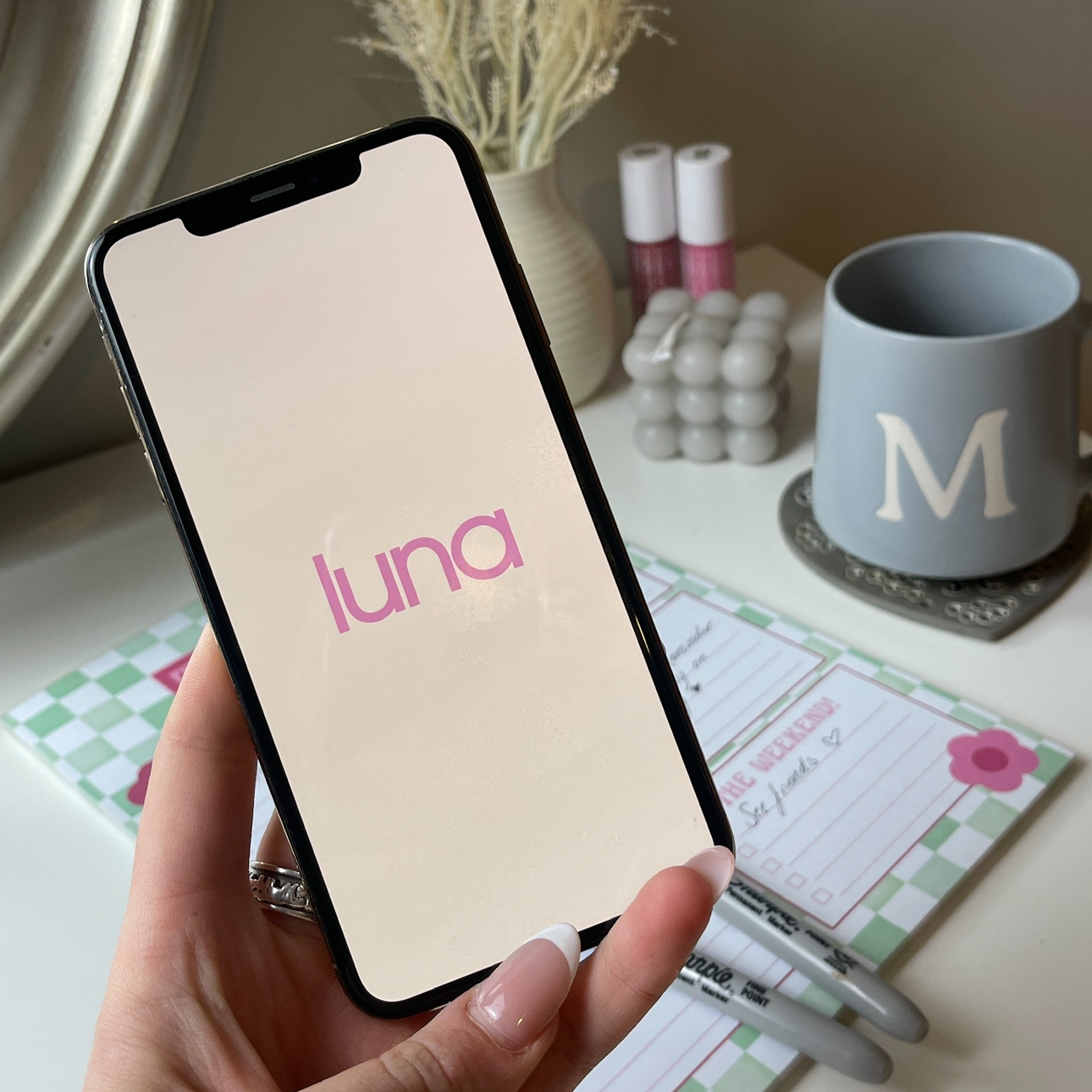 Hand holding a phone showing the luna app's loading screen, with a mug, lip products and a desk planner in the background.