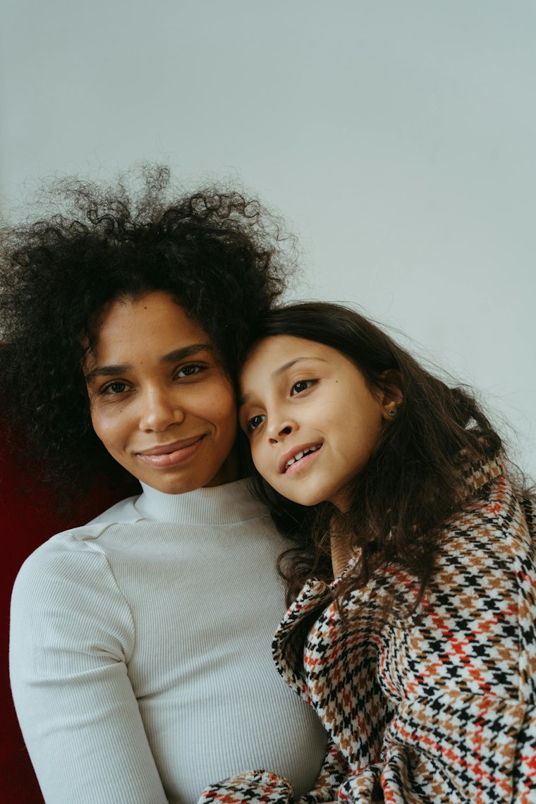 Mother and young daughter smiling closely together, with the mother in a white top and the daughter in a houndstooth coat.