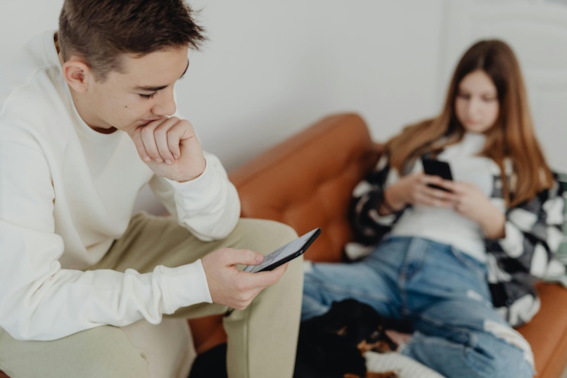 Teenage boy and girl sitting on a sofa each looking at their own phones.