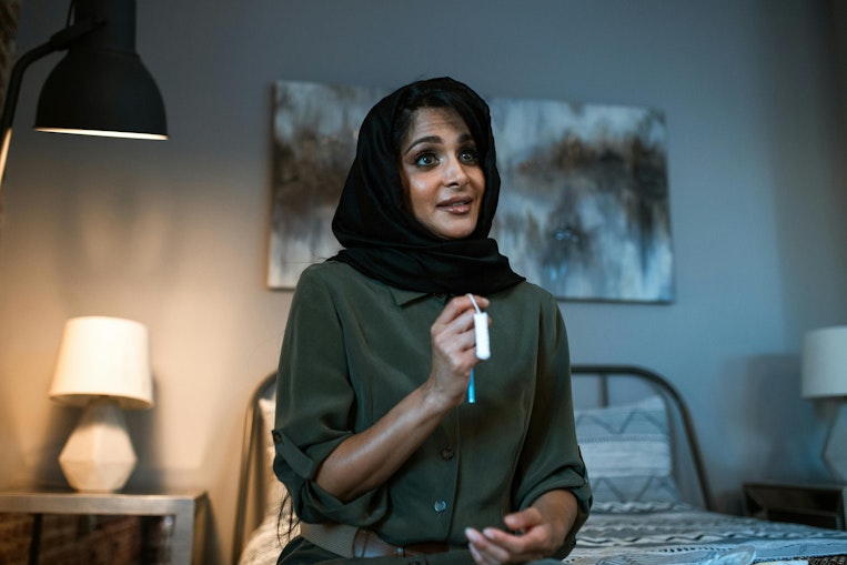 Woman in a headscarf sitting on a bed holding a tampon while talking, with lamps and artwork in the background.