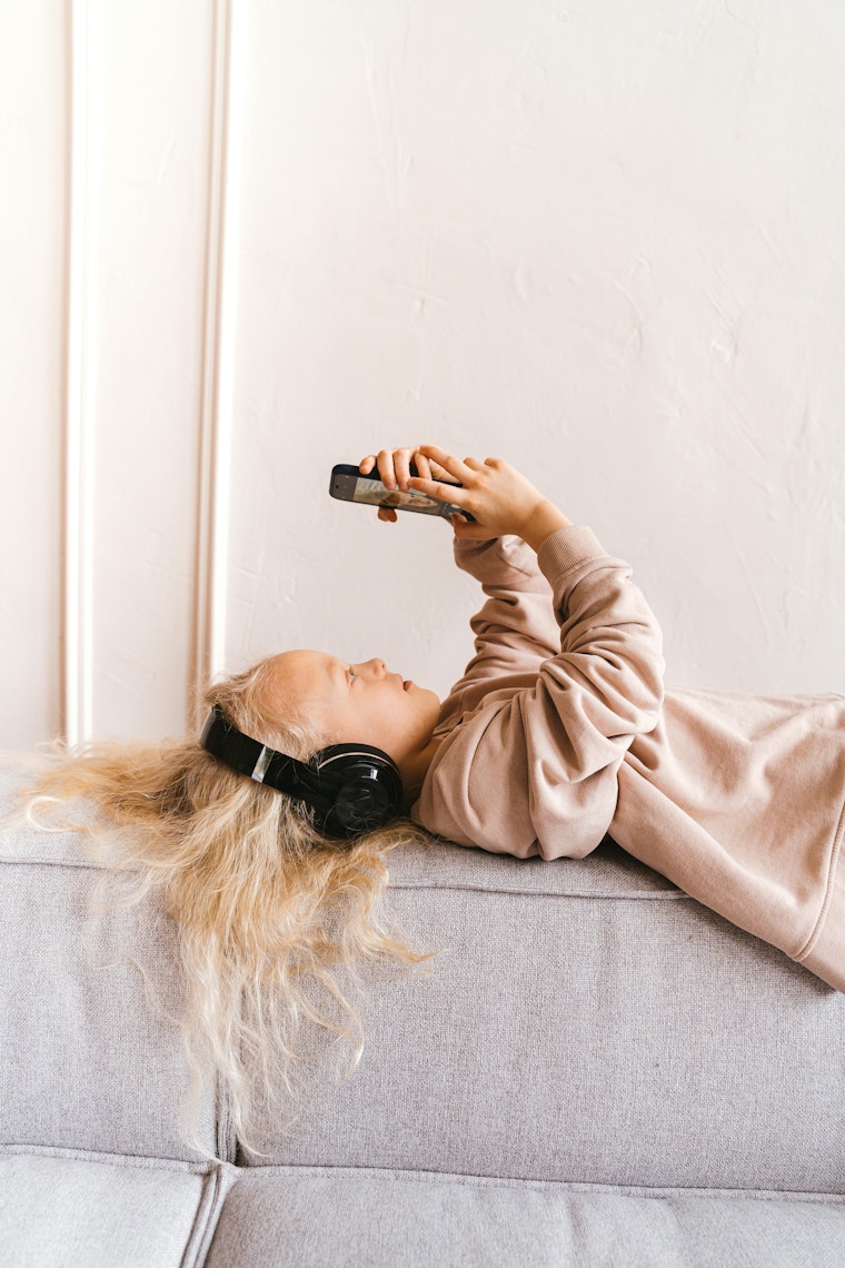 Girl lying on a sofa wearing headphones and holding up her phone.