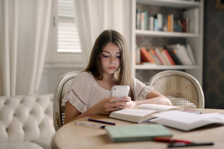 Teenage girl sitting at a table with open books and a notebook, looking down at her phone, with a bookshelf in the background.
