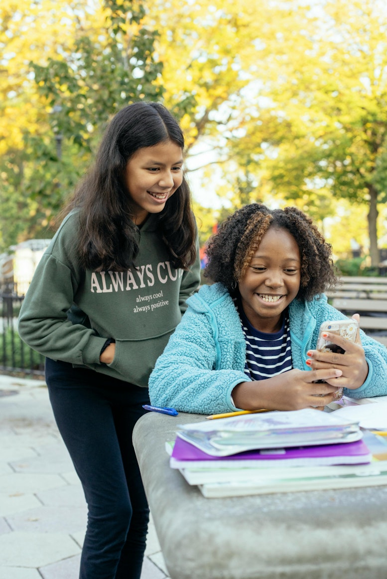 Two teenage girls laughing together while looking at a phone outdoors, with books on a table beside them.