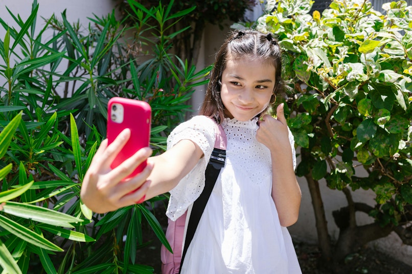 Girl in a white dress taking a selfie with a pink phone while giving a thumbs up, surrounded by greenery.