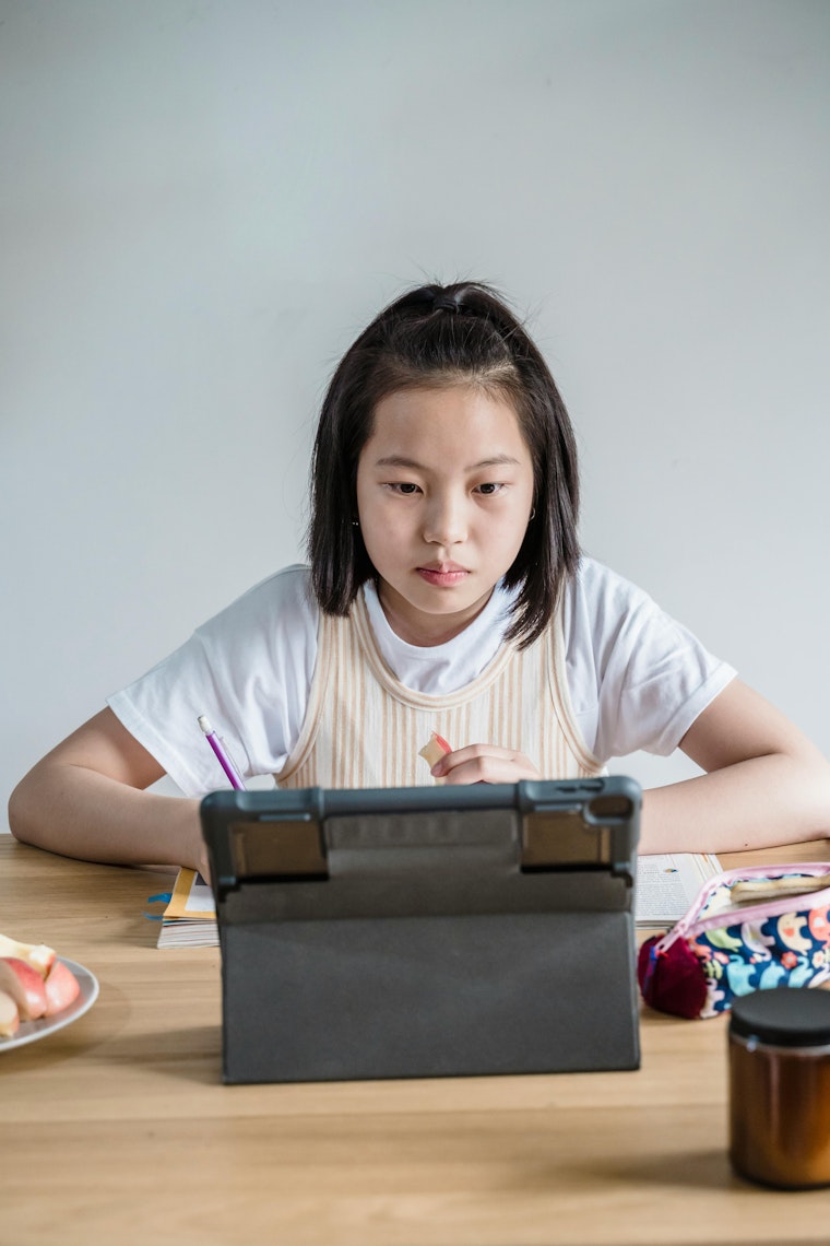 Girl sitting at a desk looking at a tablet, with a pencil case, books and a small plate of fruit nearby.