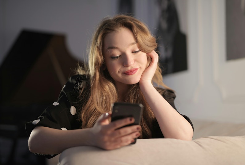 Teenage girl smiling and looking at her phone while resting her head on her hand on a sofa.