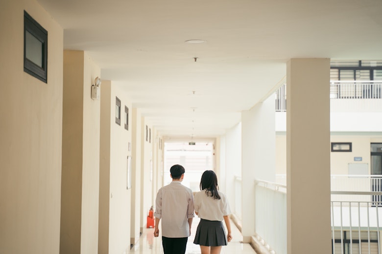 Teenage boy and girl in school uniform walking side by side down a school corridor, seen from behind.