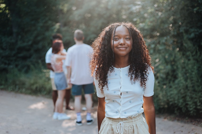 Smiling teenage girl with long curly hair standing outdoors on a path, with friends chatting in the background.
