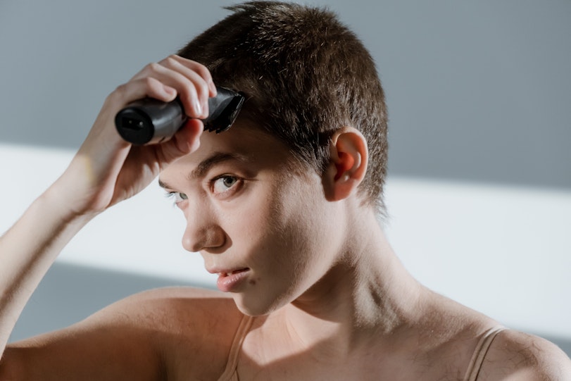 Close-up of a young person using electric clippers to shave their own hair, looking towards the camera.
