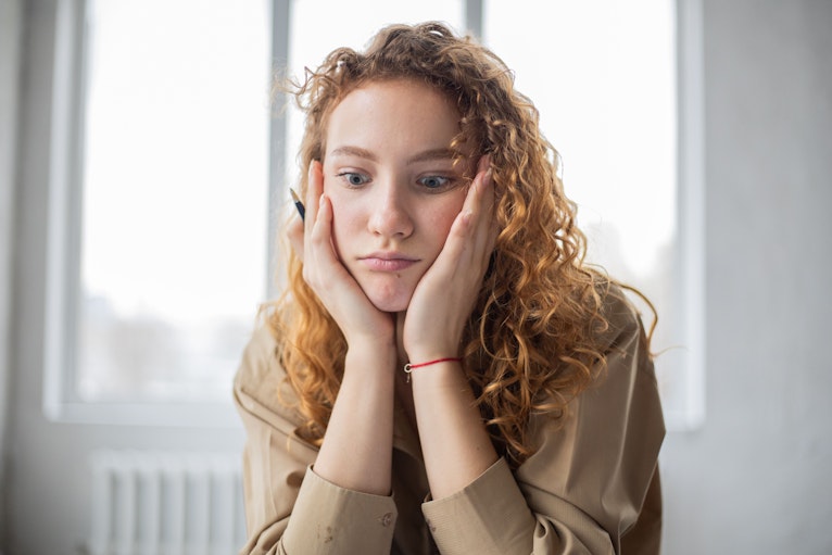 Teenage girl with curly hair resting her face in her hands, looking down with an upset expression.