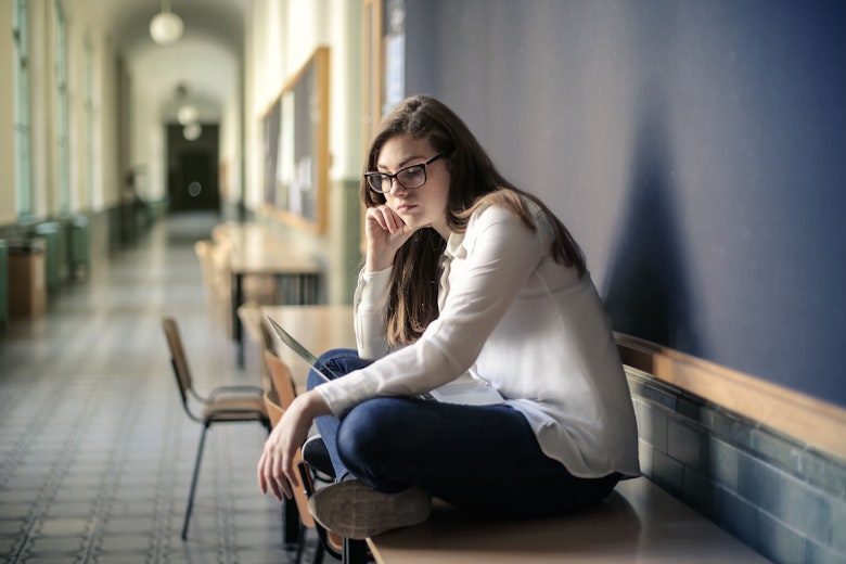 Teenage girl in glasses sitting alone on a bench in a school corridor, looking down at a laptop with a thoughtful expression.