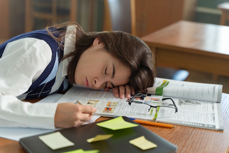 Teenage girl in school uniform asleep with her head resting on an open textbook at her desk.