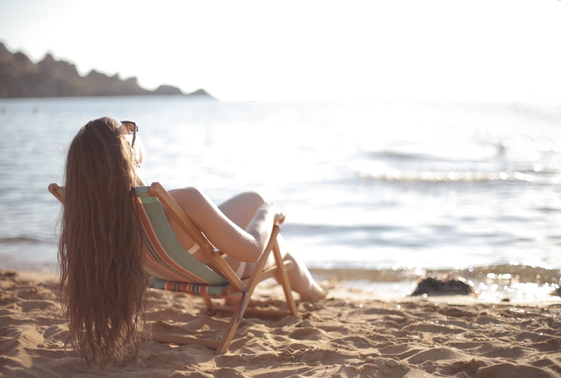 Woman with long hair reclining in a deckchair on a sandy beach, facing the sea.