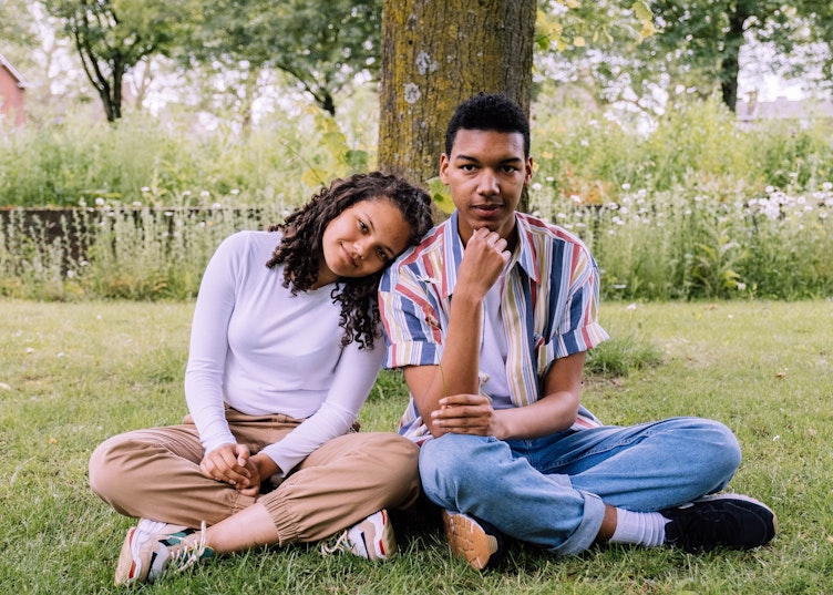 Teenage girl resting her head on a teenage boy's shoulder while they sit together on the grass under a tree, holding a small flower.