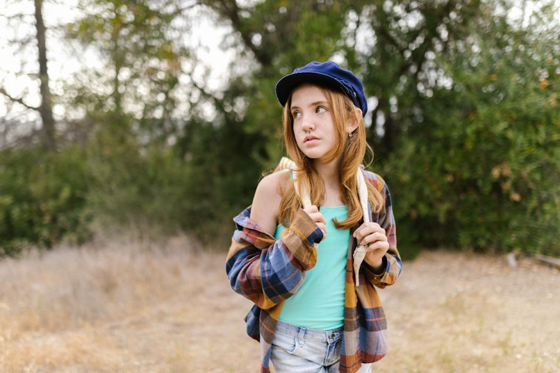 Teenage girl in a navy cap and plaid jacket over a mint tank top, standing outdoors in a field looking over her shoulder.