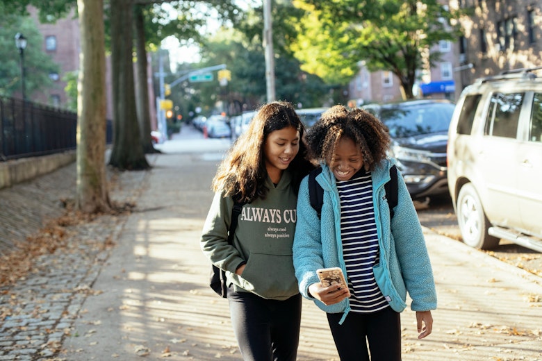 Two teenage girls with backpacks walking down a tree-lined street, smiling and looking at a phone together.