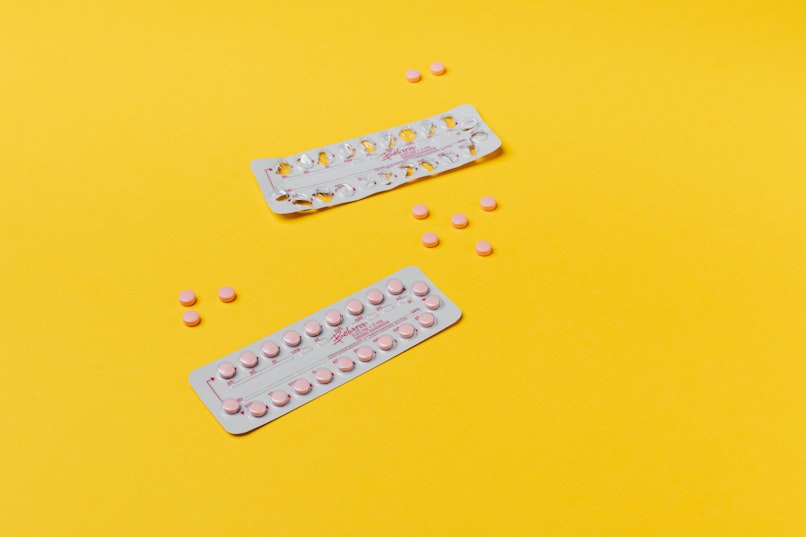 Two blister packs of pink contraceptive pills with loose pills scattered nearby on a yellow background.