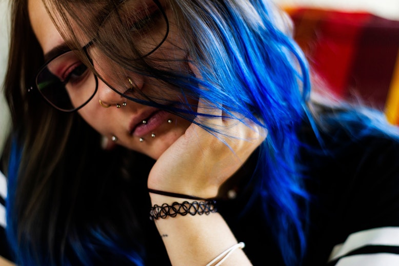 Close-up of a teen with glasses, blue-dyed hair, and multiple facial piercings including nose and lip piercings.