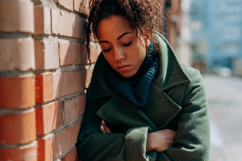Teenage girl in a green coat and scarf leaning against a brick wall with her arms crossed, looking down with a withdrawn expression.