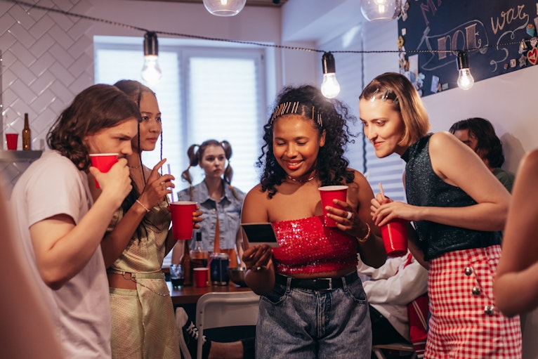 Group of teenagers at a house party holding red plastic cups, with string lights and a beer bottle visible in the background.