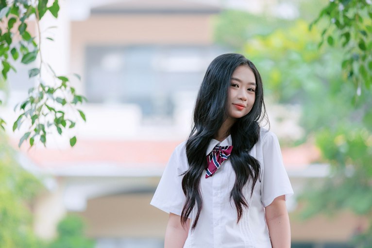 Teenage girl with long dark hair in a white school uniform with a striped neck tie, smiling outdoors with greenery behind her.