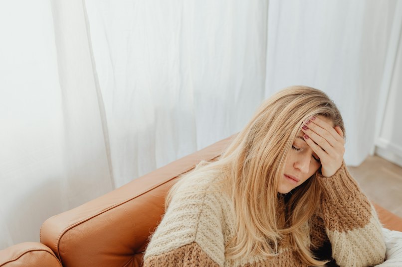 Woman sitting on a couch with her hand pressed to her forehead, looking tired and uncomfortable, in front of sheer white curtains.