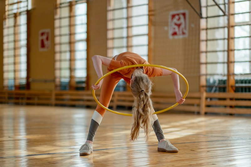 Teenage girl in orange sportswear performing a rhythmic gymnastics move with a yellow hoop in a school gym.