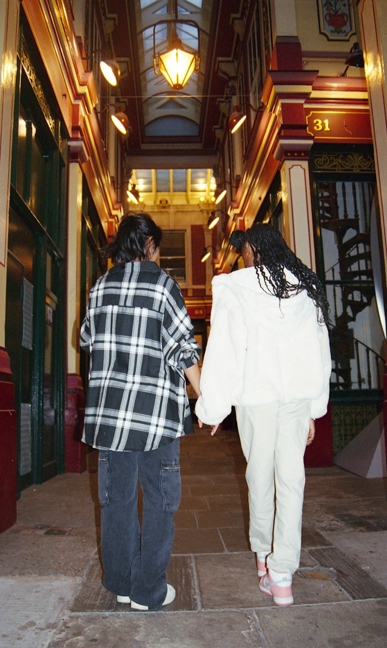 Two teenage girls walking hand in hand through a covered Victorian arcade with glowing lanterns, one in a checked shirt and baggy jeans and the other in a white fluffy jacket and cream trousers.