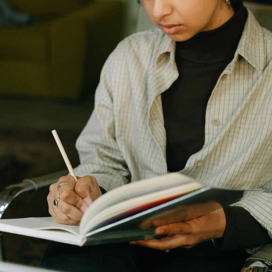 Person in a striped shirt and black polo neck writing in an open notebook with a pencil.