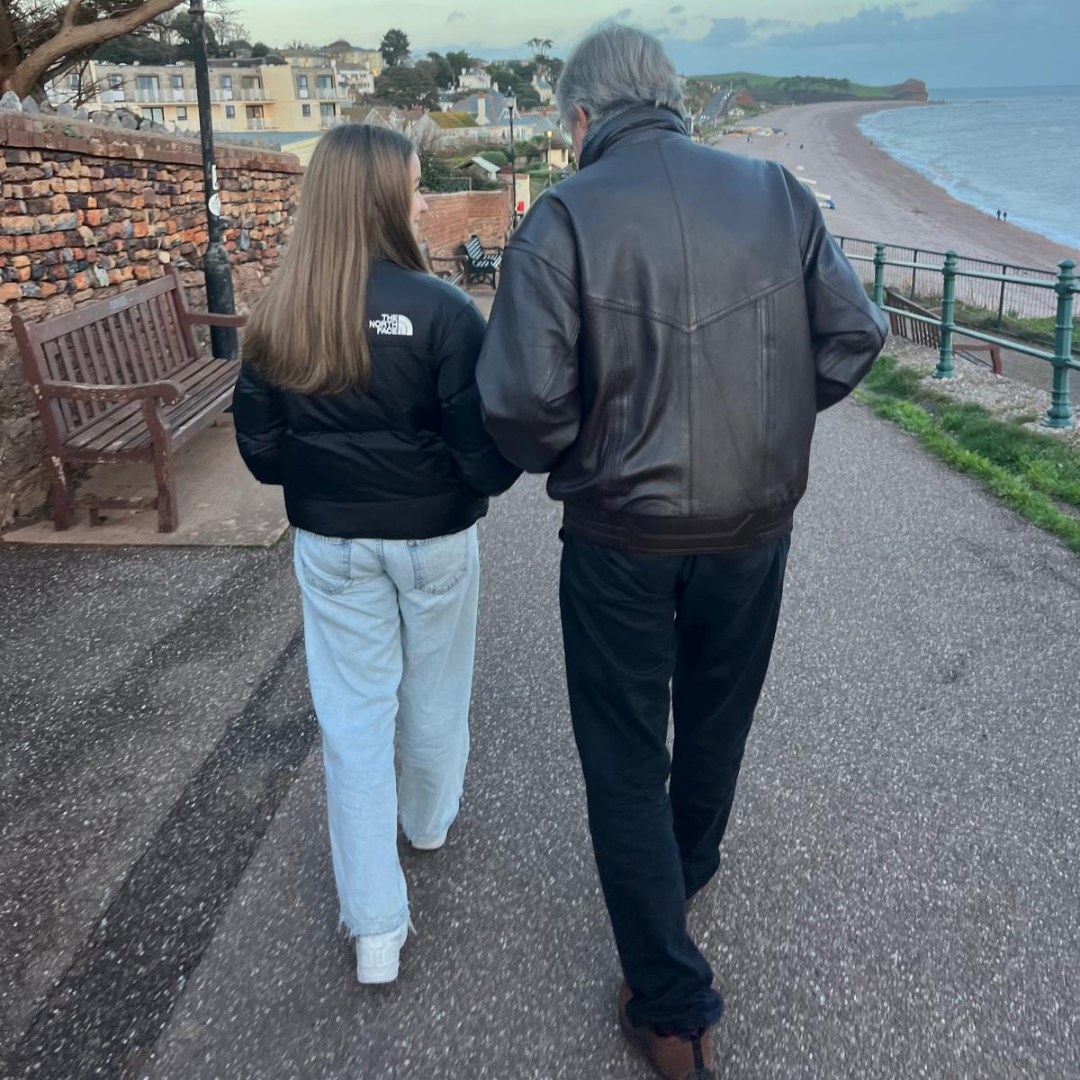 Teenage girl with long hair in a black puffer jacket and light jeans walking arm in arm with a grey-haired man in a leather jacket along a seafront path overlooking a beach and red cliffs.
