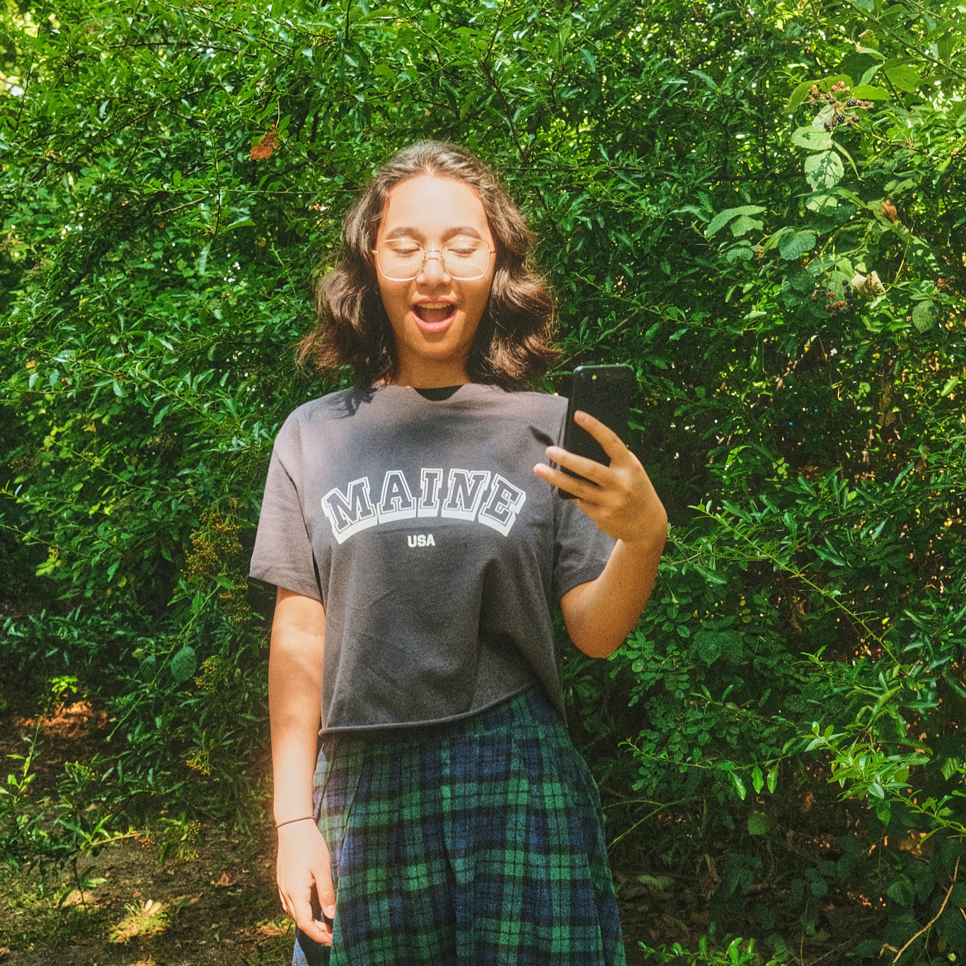 Teenage girl with wavy hair and glasses looking excitedly at her phone, wearing a grey “Maine USA” T-shirt and a green tartan skirt in front of leafy green bushes.