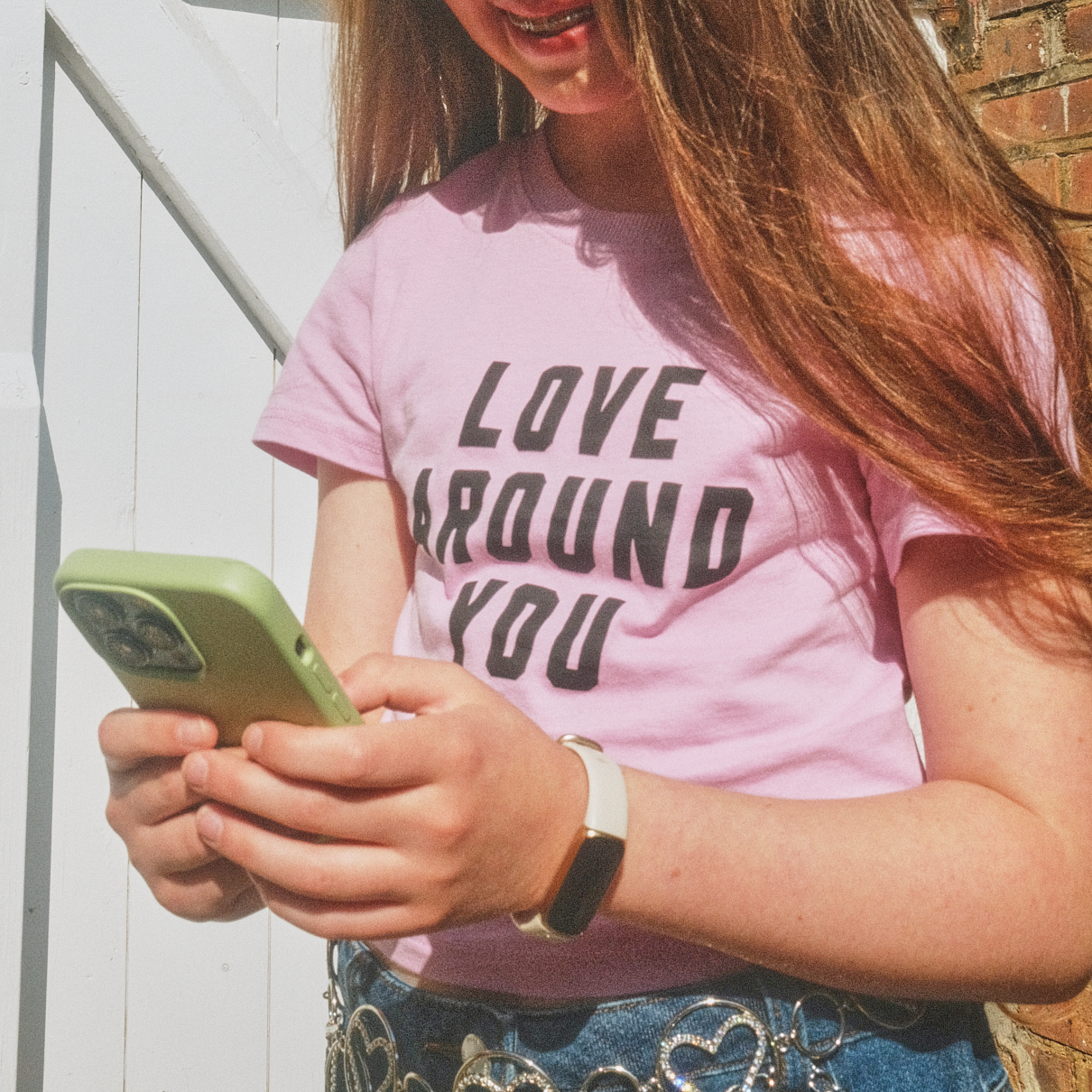Teenage girl in a pink “Love around you” T-shirt, jeans and a heart chain belt, typing on a green phone outside by a white door.