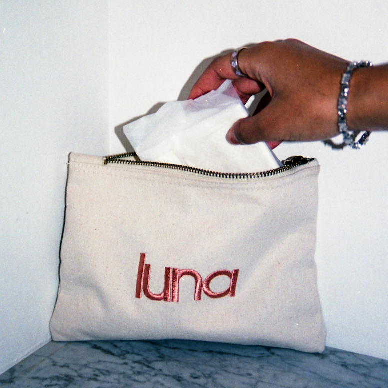 Hand with a silver bracelet and ring pulling a wrapped period pad out of a cream canvas pouch with “luna” embroidered in pink.