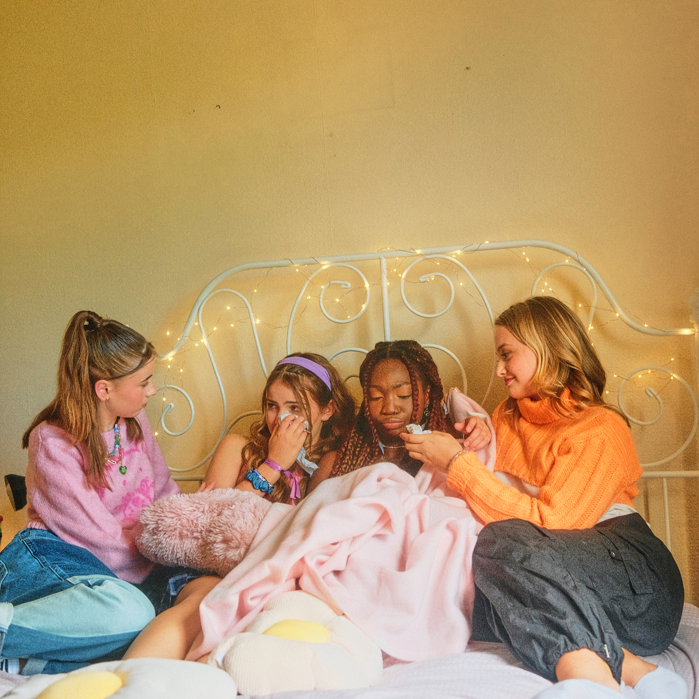 Four teenage girls sitting together on a bed under fairy lights, with friends comforting a girl who is wiping her eyes with a tissue.