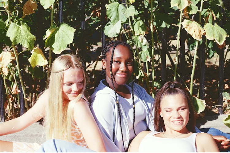 Three smiling teenage girls sitting together in the sunshine in front of railings covered in large green leaves.