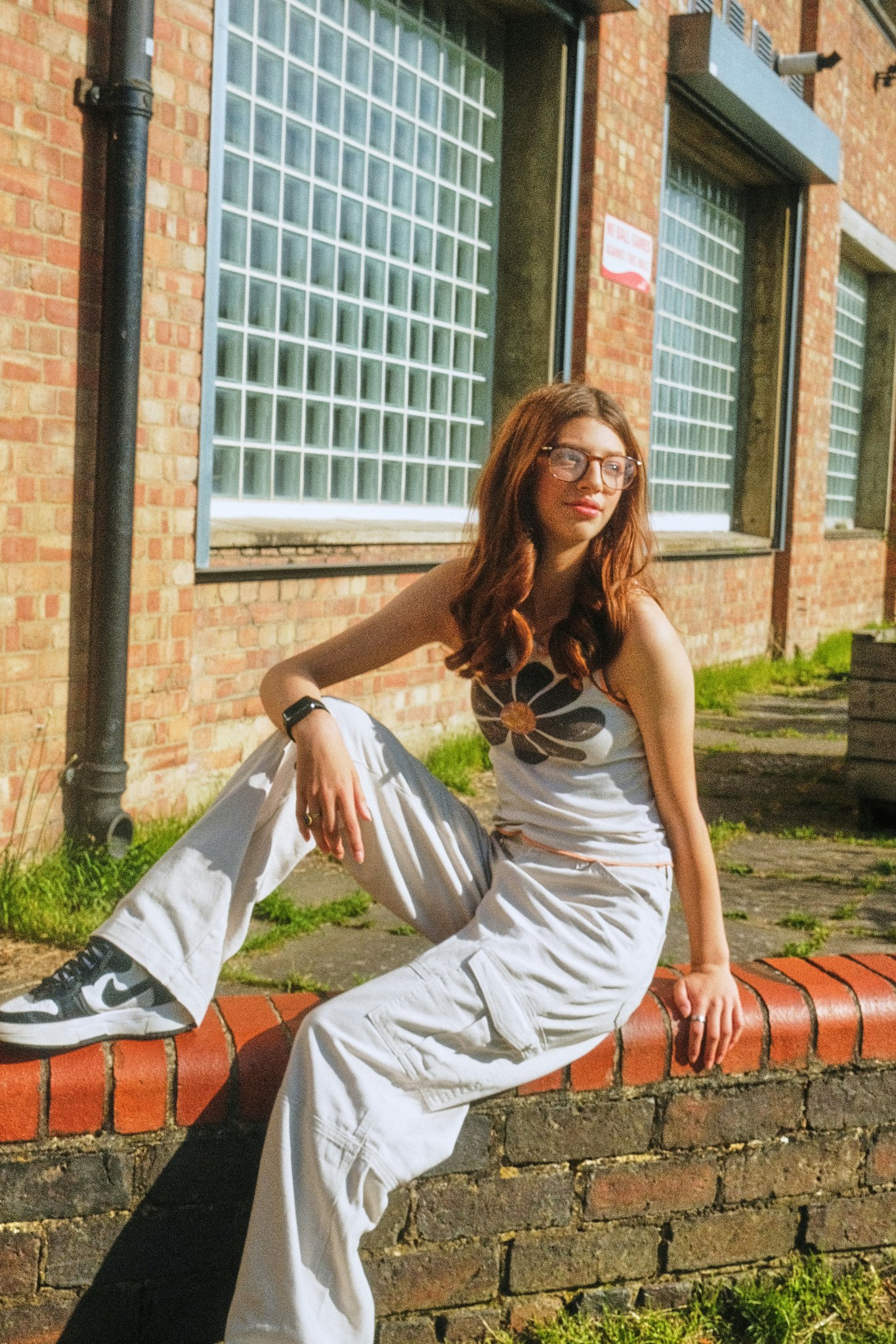 Teenage girl with long auburn hair and glasses sitting on a low brick wall in the sunshine, wearing a flower-print top and light cargo trousers.