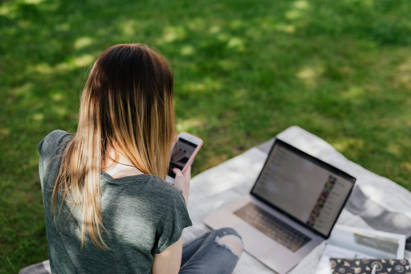 Girl with long hair sitting on a blanket on the grass, looking at her phone next to an open laptop.