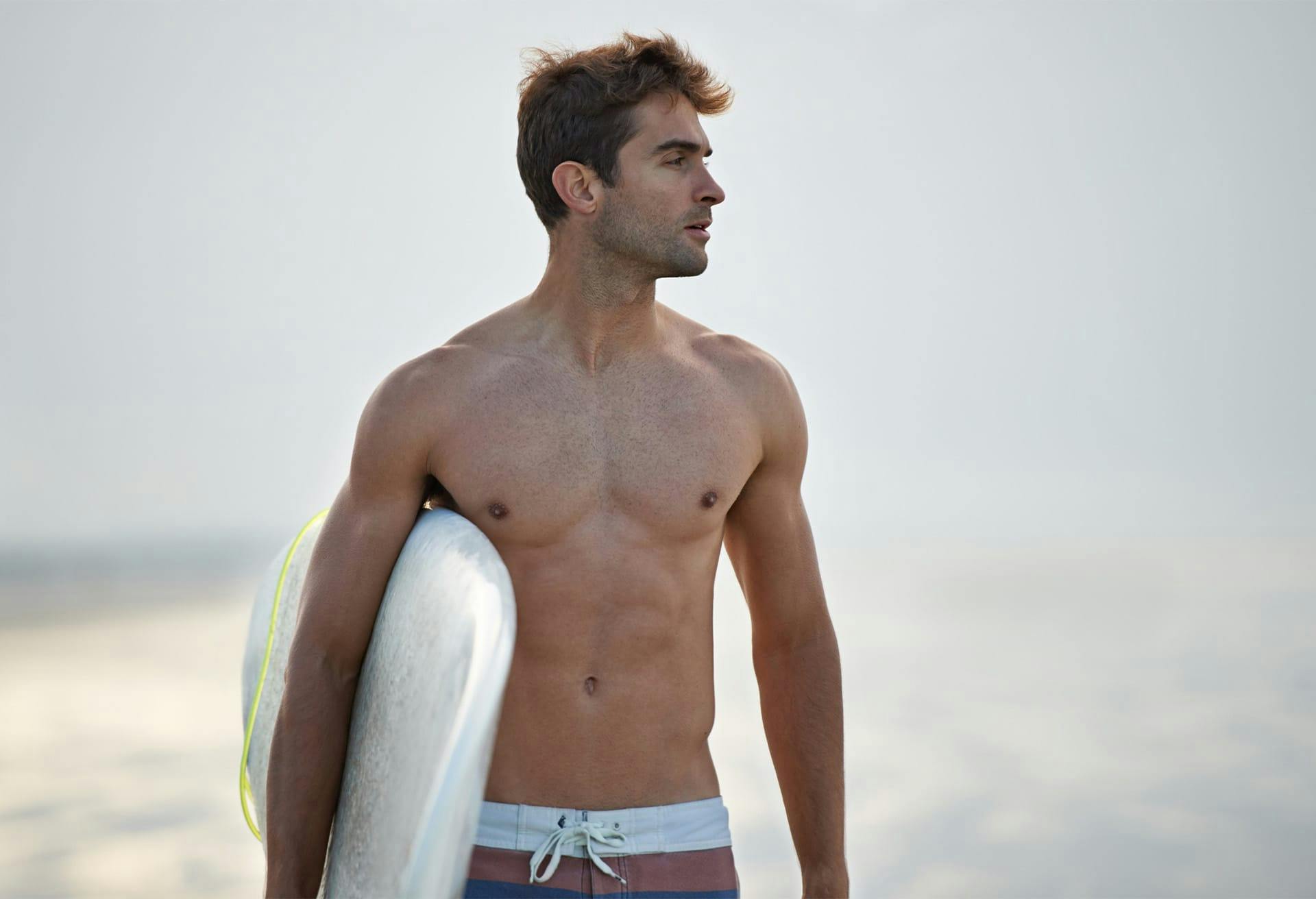 Man walking shirtless with surfboard on beach