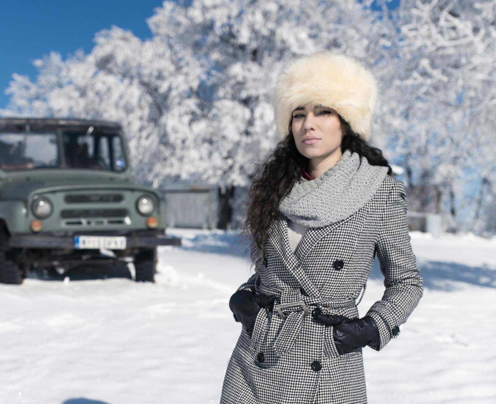 woman in a coat and hat standing in the snow with a jeep behind her