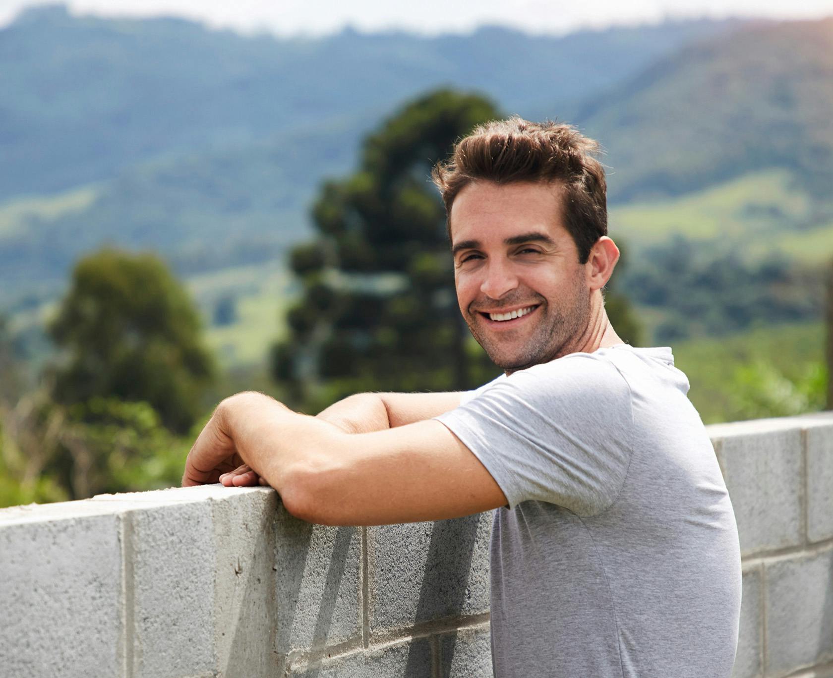 smiling man leaning on a wall with mountains in the background
